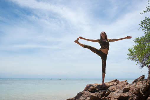 A young woman visiting Thailand does the Extended Hand to Big Toe Yoga Pose (Utthita hasta Padangusthasana) at a beach on Ko Phangan, Thailand