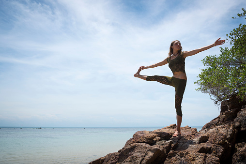 A young woman visiting Thailand does the Extended Hand to Big Toe Yoga Pose (Utthita hasta Padangusthasana) at a beach on Ko Phangan, Thailand