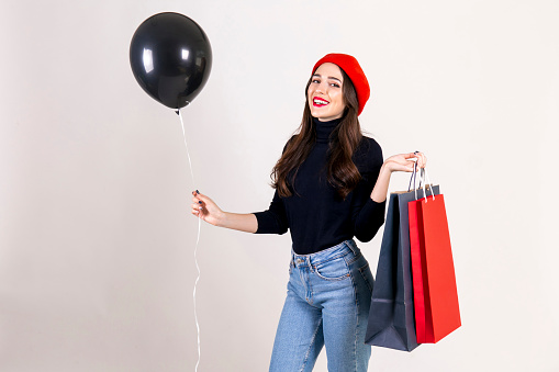 Beautiful hipster young woman with bright red lipstick, wearing turtleneck, mom jeans, red beret, holding blank balloon and colorful shopping bags. Black friday sale concept. Background, copy space.