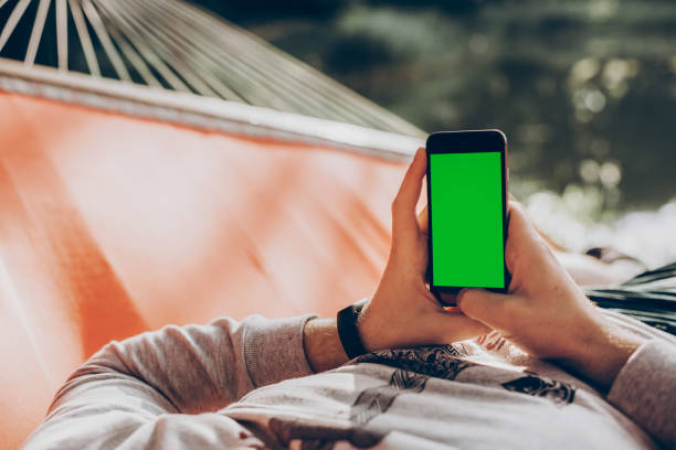 istock hipster man holding smart phone with empty screen with space for text, and relaxing in hammock in sunny summer park. mock up. guy looking at blank phone, social media concept 1055635562