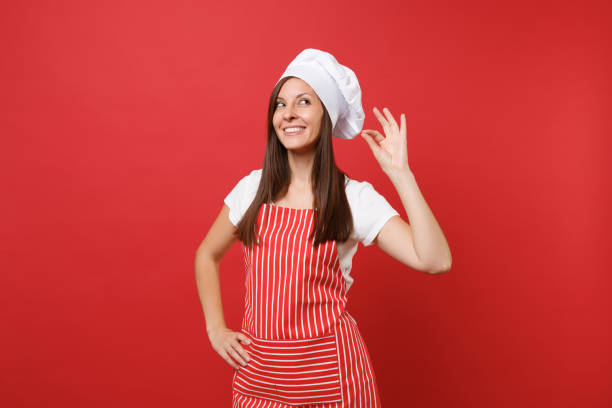 主婦女性シェフや縞のエプロンでパン屋は白い t シャツ、赤い壁の背景に分離されたトーク シェフ帽子です。穏やかなかわいい女性をいい味喜び記号を笑っています。コピー スペース コン� - hat women chef occupation ストックフォトと画像