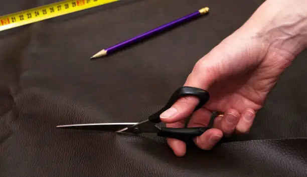 Woman hand cutting out brown leather with scissors. Purple pencil and yellow tape measure. Concept of cutting-out and sewing