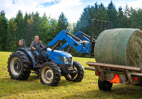 A farmer on a tractor loading hay bales on a trailer in a field