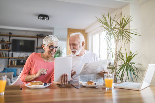 Modern senior couple looking at newspaper and a tablet during breakfast Cute and lovely senior adult couple, spending their time together in love and tranquility, enjoying a morning breakfast. human settlement stock pictures, royalty-free photos & images