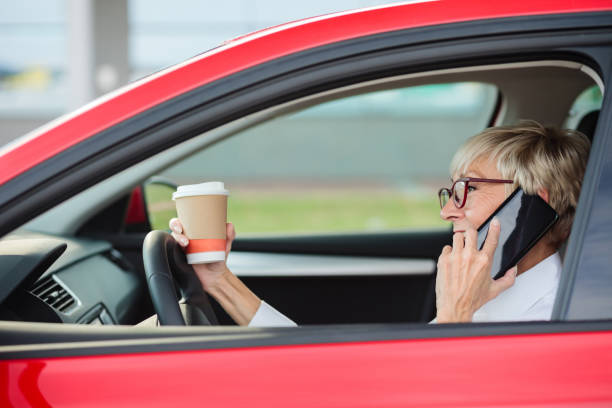 imprudente, sonriente mujer madura hablando por teléfono y sosteniendo una taza de café mientras se conduce un coche - driving car distracted accident fotografías e imágenes de stock