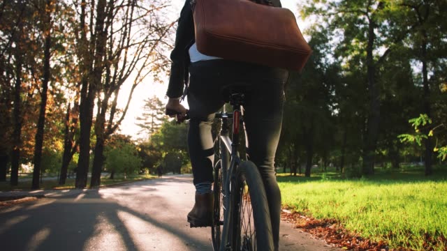 Young businessman with leather bag riding bicycle to city park, slow motion, back view