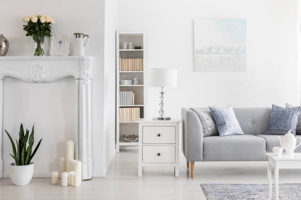 istock Lamp on cabinet and bookshelf next to grey sofa in white flat interior with painting and flowers. Real photo 1055867340