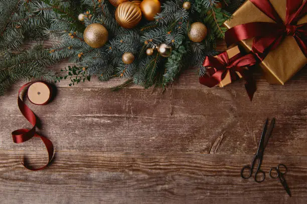 Photo of top view of Christmas gift boxes, fir wreath, ribbon spool and scissors on wooden table