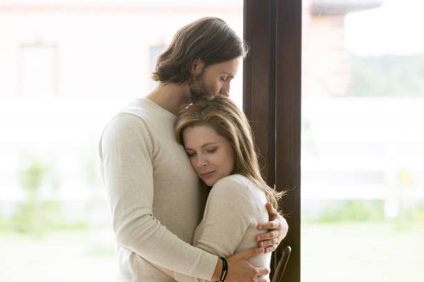 Sad young married couple embracing standing in living room Sad young married couple embracing standing in living room opposite window at home. Sorrowful wife and husband feels unhappy, thinking about problems in relations, miscarriage, misbirth or infertility arguing couple divorce family stock pictures, royalty-free photos & images