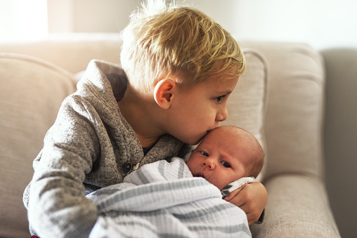 Shot of a cheerful little boy holding his little infant brother and giving him a kiss on the forehead while being seated on a sofa at home during the day