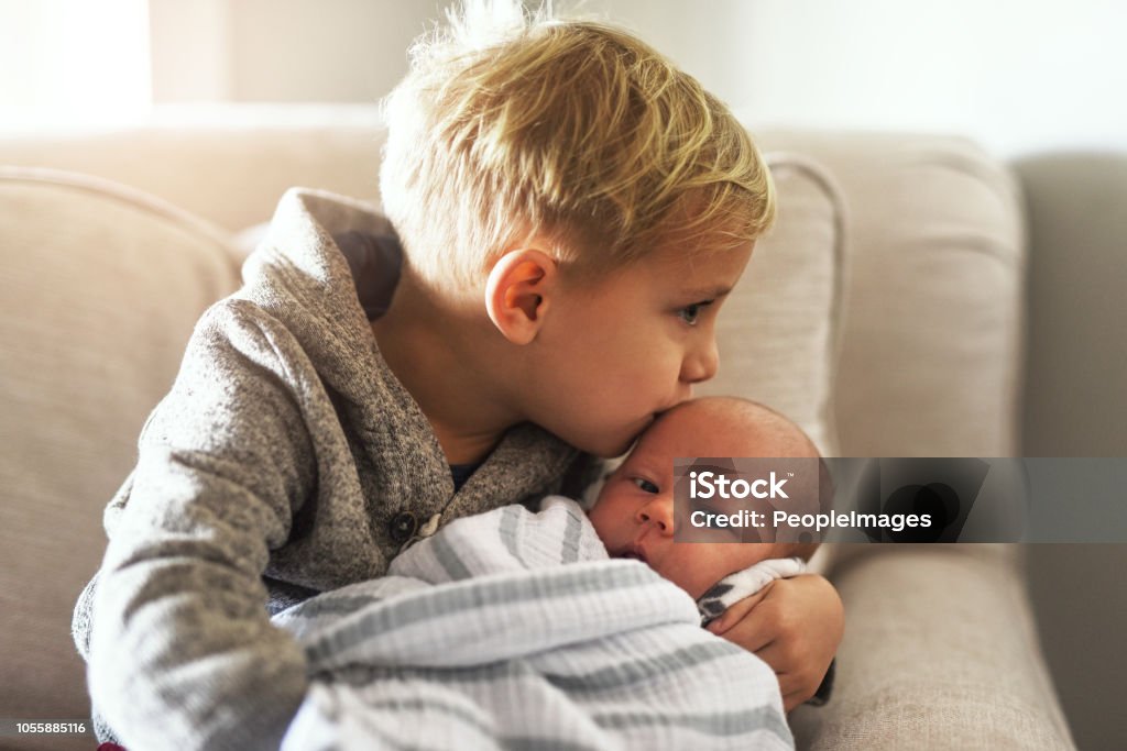 Too much cuteness! Shot of a cheerful little boy holding his little infant brother and giving him a kiss on the forehead while being seated on a sofa at home during the day Sibling Stock Photo