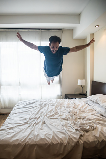 Happy young man on vacations jumping on bed looking very excited and smiling