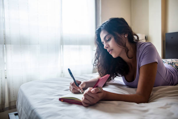 istock Beautiful woman lying down on her bed writing on her diary looking thoughtful 1055888684