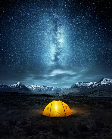 Camping in the wilderness. A pitched tent under the glowing  night sky stars of the milky way with snowy mountains in the background. Nature landscape photo composite.