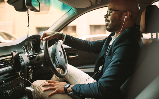 Side view of young man in suit listening music while driving car. African businessman driving a vehicle to office.