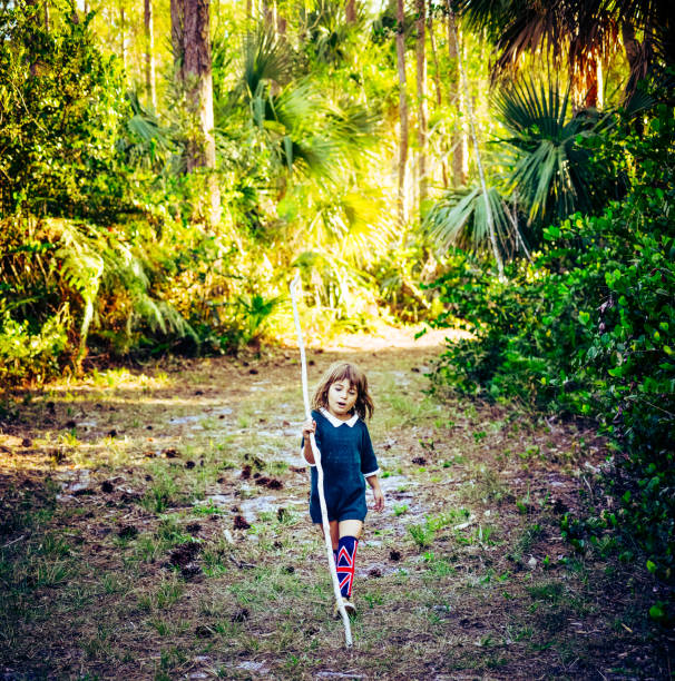 Child, little girl, carries a huge walking stick in a rural urban, woodsy forest. She is smiling, happy, cheerful. Hiking, or walking, using her imagination, enjoying nature and the wilderness. She wears a retro vintage outfit and British flag knee socks