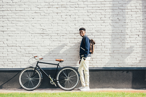 cheerful stylish african american man with leather backpack standing with bicycle near white brick wall