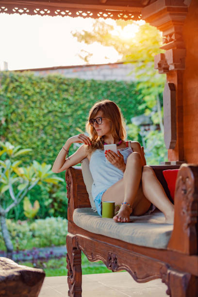 istock Young woman using cellphone and drinking coffee / tea on a porch sofa. 1055937700