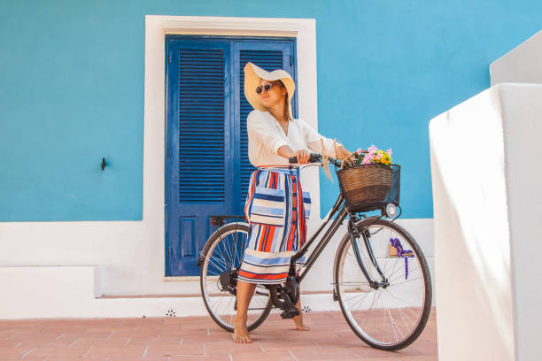 ポンツァ島イタリアの日当たりの良い夏の日、大きな帽子とカラフルなファッションのドレスを着て青い家の前に自転車を持つ美しい少女 - italy house sunny outdoors ストックフォトと画像