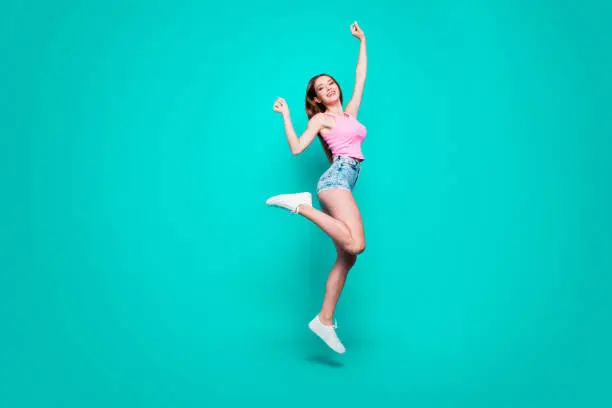 Photo of Full size body length of young slim magnificent sweet laughing girl in top and jeans shorts, sneakers, dancing in air, raising hands up, celebrating. Isolated over bright vivid turquoise background