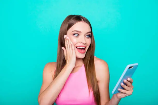 Photo of Portrait of young perfect straight-haired cheerful amazed girl wearing casual pink shirt, reading winning sms. Isolated over bright vivid turquoise teal background