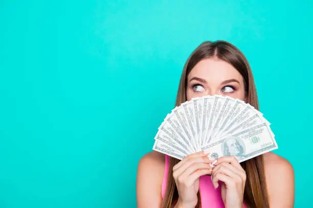 Photo of Attractive gorgeous young amazed girl wearing pink blouse, excited, covering face with dollar banknotes. Copy space. Isolated over bright vivid blue teal, turquoise background