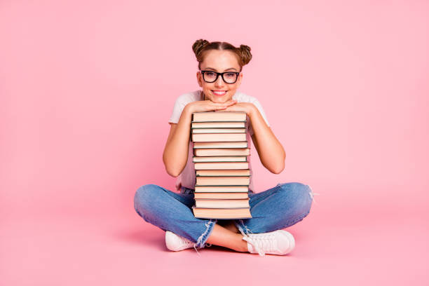 pernas completa, corpo, retrato de tamanho da menina de leitor em jeans azul, camiseta branca e tênis se senta no chão, no livro de porões de sala de aula e com alegria, olha para a câmera isolada no fundo rosa vívido - nerd student female exam - fotografias e filmes do acervo