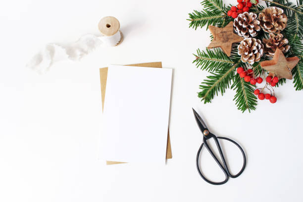 Christmas composition. Blank greeting card, envelope mock-up scene. Decorative corner. Pine cones, fir tree branches, red rowan berries and wooden stars on white table background. Flat lay, top view.