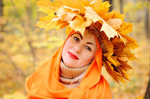 A large portrait of a beautiful middle-aged blonde in a wreath of autumn leaves, which stands in an orange scarf in the forest on a yellow background, looks at the camera and smiles. Plus size woman.