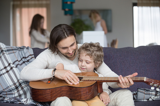 Family with children at home. Little preschool son sits on loving father lap on couch in living room, mother and daughter out of focus on background. Patient male learning teaching how to play guitar