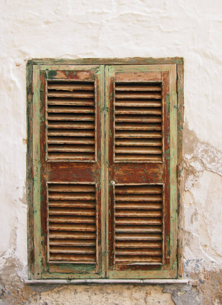 ancient closed green painted brown window shutters in a white wall with cracked cement and plaster