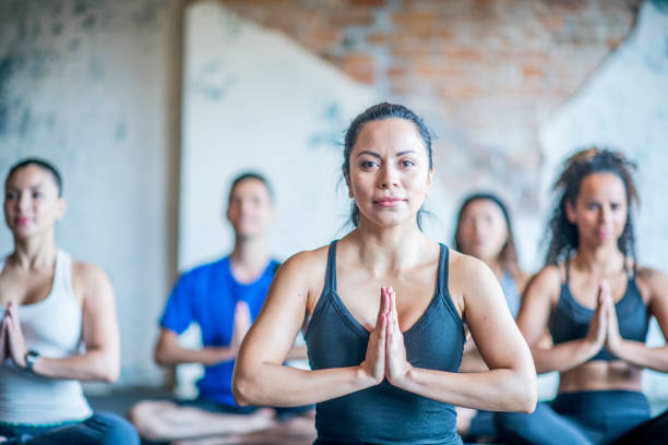 Young woman sitting and meditating in lotus pose with group. Group of fit females meditating in a gym class.