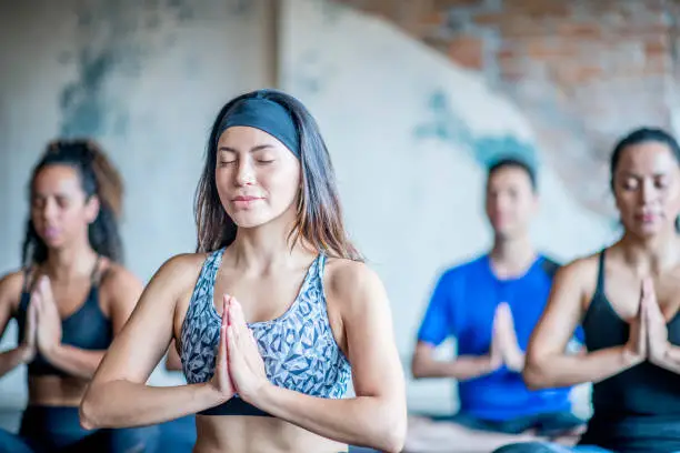 Young hispanic women wearing headband sits in yoga pose with her eyes closed. She looks relaxed and at peace. She is in a group workout.