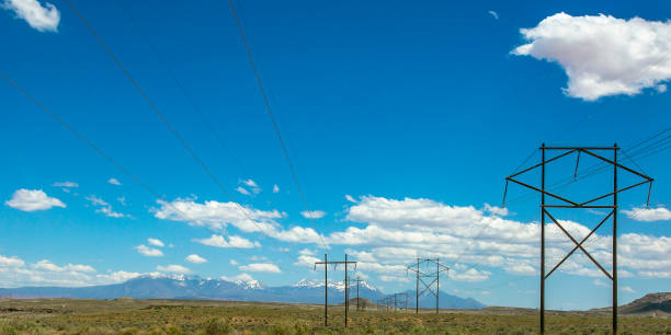 Power lines with view of mountain under blue sky. Power lines on a vast grassy field in Moab, Utah. A white-peaked mountain looks majestic in the distance beneath brilliant blue sky with puffy clouds.