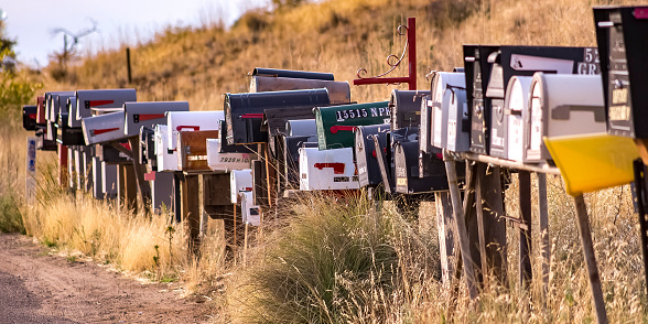 Row of mailboxes on a grassy roadside. Metal mailboxes placed in a row on a grassy ground beside a road. The grassy terrain continues into a slope behind the sunlit mailboxes.