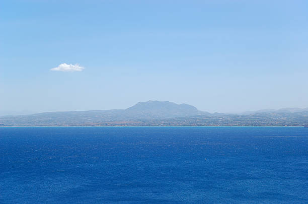 A serene coastal landscape featuring a rocky shoreline, calm sea, and distant hills under a cloudy sky. The tranquil scene captures the natural beauty of the seaside