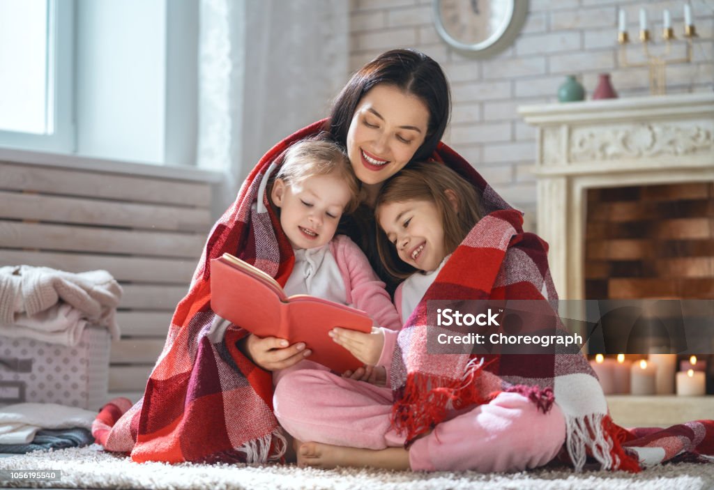 mother reading a book to her daughters Winter portrait of happy loving family. Pretty young mother reading a book to her daughters at home. Winter Stock Photo