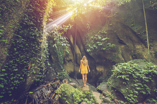 Girl in yellow dress standing in tropical rainforest cave looking up at the magnificent sunbeam coming from the rocks above. People travel wonderlust nature concept