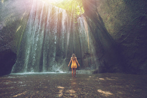 Girl standing in tropical rainforest cave looking up at the magnificent sunbeam coming from the rocks above. People travel wonderlust nature concept