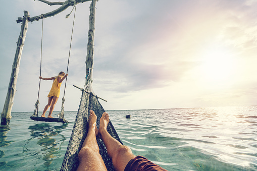 Young couple swinging on the beach by the sea, beautiful and idyllic landscape. People travel romance vacations concept. Personal perspective of man on sea hammock and girlfriend on sea swing.
