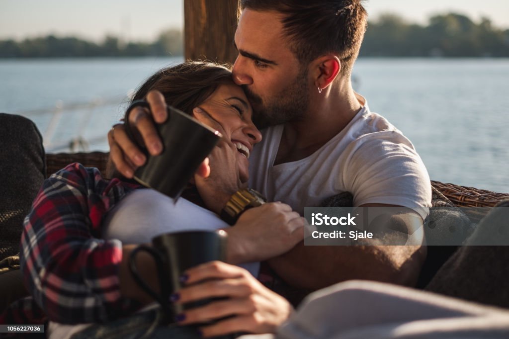 Boyfriend kissing his smiling girlfriend in forehead by the river Boyfriend kissing his smiling girlfriend in forehead while she is lying covered with blanket  by the river. Beautiful autumn day Couple - Relationship Stock Photo