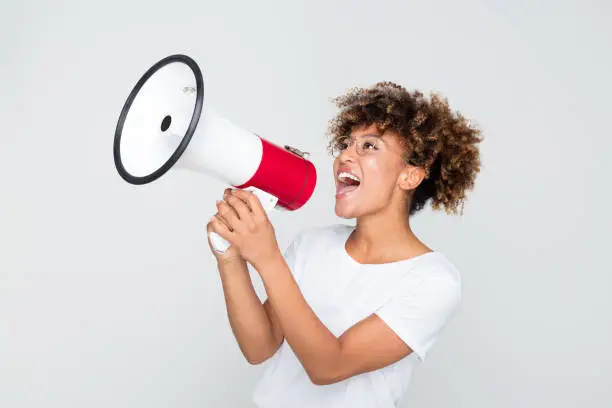 Portrait of young afro american woman yelling into a megaphone on grey background. African female making an announcement with megaphone.