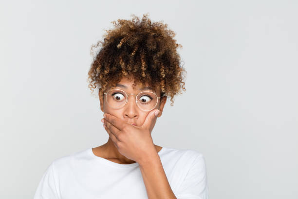 Terrified afro american woman Close up of terrified afro american woman looking away with hand on mouth against grey background. Female with curly hair wearing eyeglasses looking terrified. staring stock pictures, royalty-free photos & images