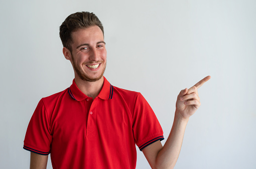 Cheerful guy showing new product, service, offer. Joyful young man in red polo pointing finger away at copy space. Presentation and promotion concept
