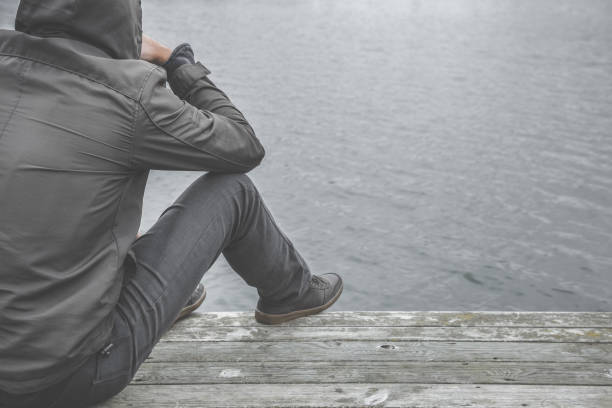 jeune homme assis sur le bord de la passerelle au lac. souffrant de difficultés de la vie, brisé le coeur ou des problèmes psychologiques. pensées sombres, sombres. isolement social. cool temps automne. - dark edge photos et images de collection