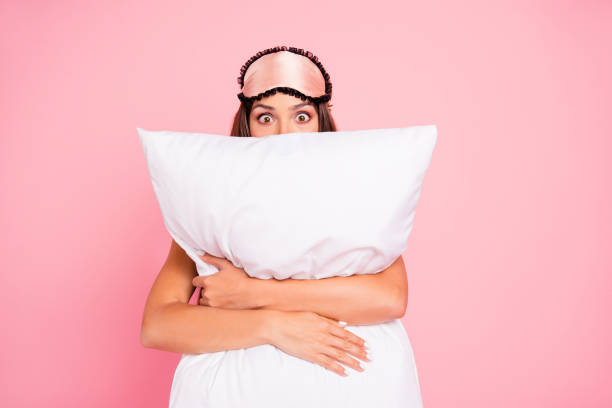 молодая великолепная шокированная дама в маске для глаз, обнимаем подушку, прячется. изолированные на розовом фоне пастели - подушка стоковые фото и изображения