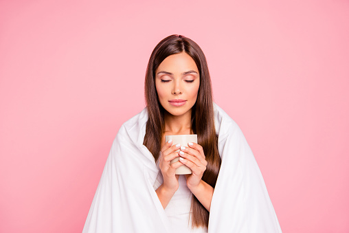 Autumn melancholy. Portrait of young smiling lady wrapped in blanket, drinking coffee. Isolated over pink pastel background