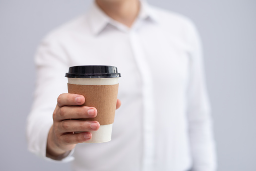 Close-up of male hand holding takeaway coffee. Young Caucasian businessman giving coffee or tea cup. Coffee break concept