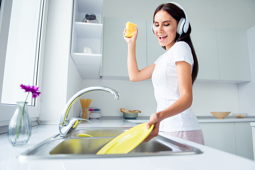 Adorable attractive young brunette smiling woman wearing pajama, washing dishes, listening to music, singing in light kitchen