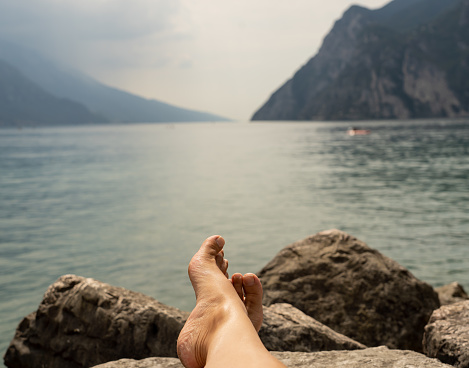 Close up feet of woman relaxing by a beautiful big lake lying on the edge of rocks on the background of mountain landscape in leisure Vacation Summer Holidays concept.